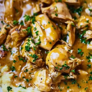 A close-up image of a dish featuring crockpot chicken with mushrooms in a creamy gravy, garnished with finely chopped parsley, served over mashed potatoes.